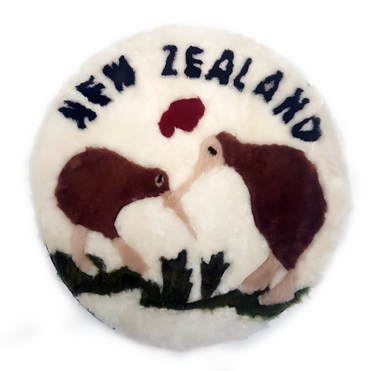 A round throw pillow with a white wool base featuring a embroidered design of two stylized kiwi fruits in a love embrace, surrounded by a green and brown border.