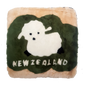 A square-shaped cushion with a green background featuring a white baby lamb on top of a grass-like pattern. The cushion also has the text 'NEW ZEALAND' at the bottom.